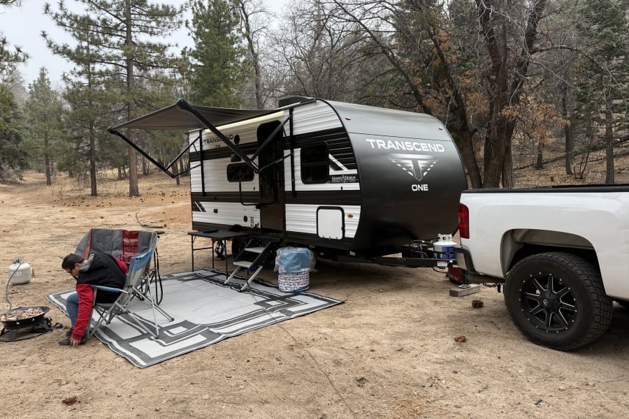 Transcend RV rental set up for camping, with a person cooking outside amidst pine trees and a white truck nearby.