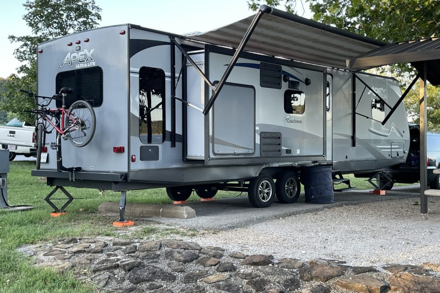A grey Apex RV rental, a travel trailer, is parked and leveled at a gravel campsite with its awning out and bike on back.