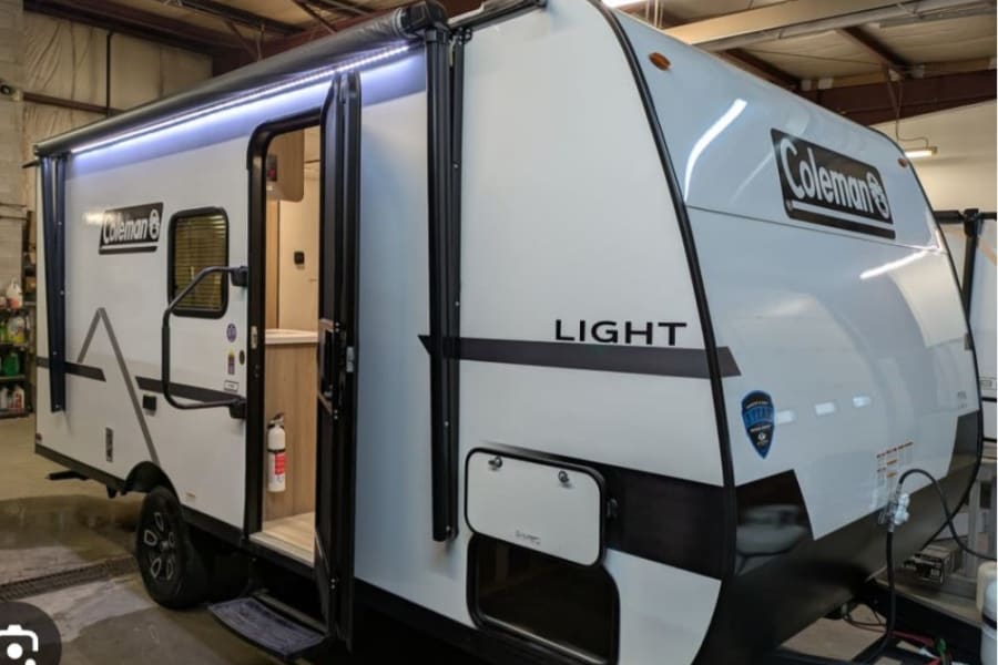 Explore with this Coleman Light travel trailer, perfect for rv rental. White with black trim, awning lights, and open door.