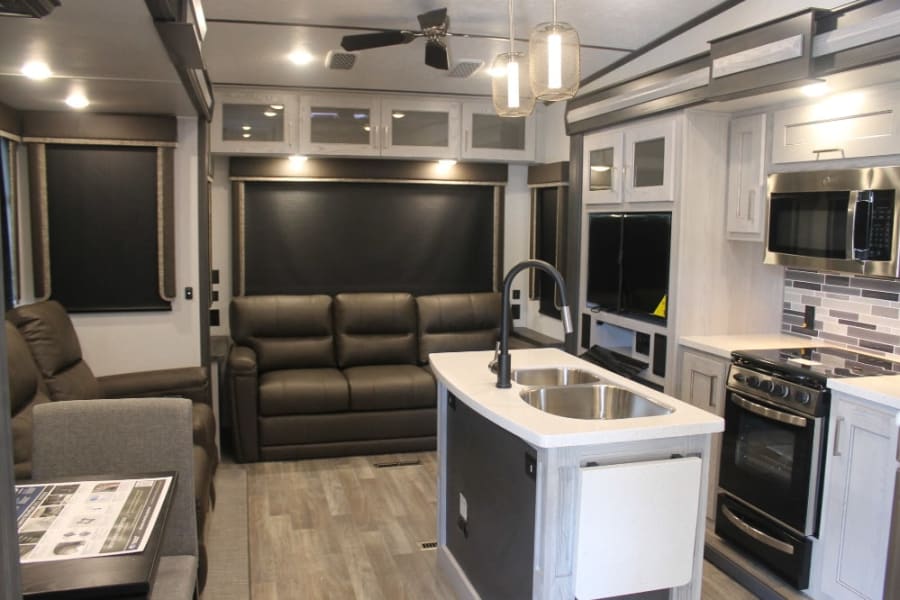 Modern RV rental interior: large living area with dark sofas, kitchen island with sink, and full kitchen appliances.