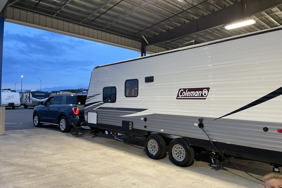 Coleman RV rental connected to a blue SUV, parked under a large covered structure at dusk.