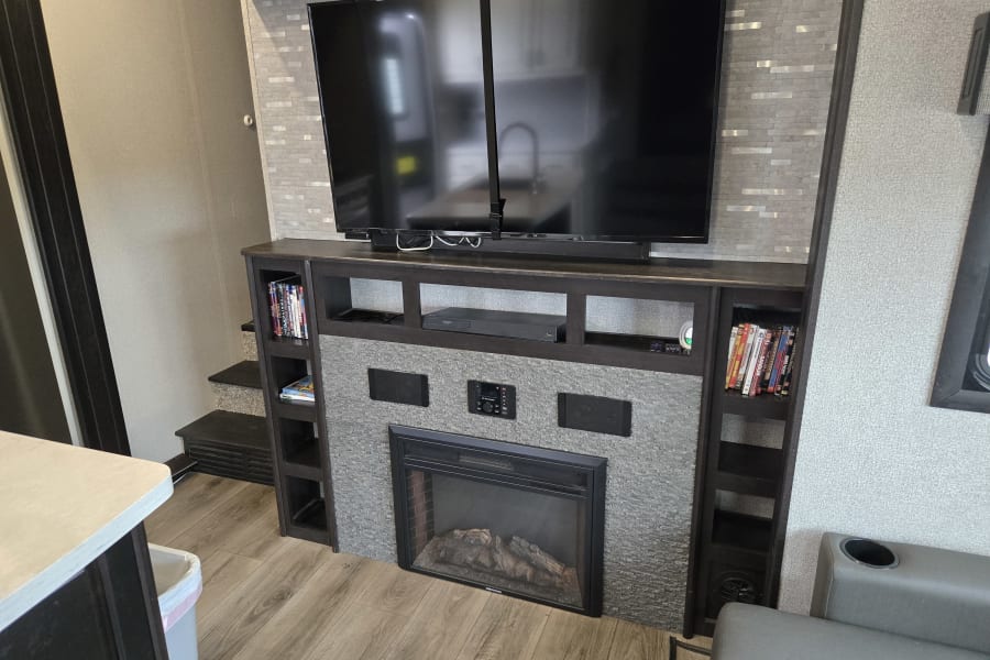 A modern RV rental living area with a dark wood entertainment center, large TV, built-in electric fireplace, and shelves.