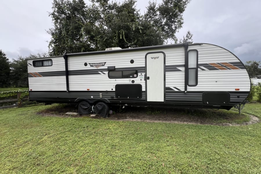This white and grey RV rental travel trailer with orange accents is parked on a grassy patch under a cloudy sky.