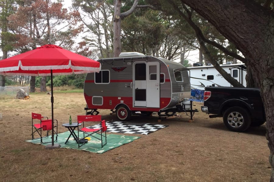 A stylish red & gray retro RV rental is set up at a leafy campsite with a red umbrella, chairs, and truck.