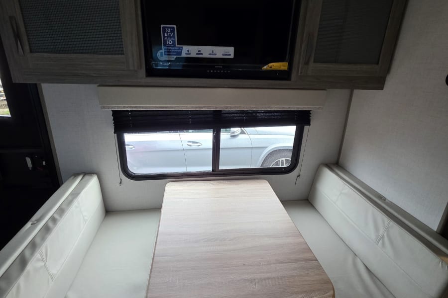 Inside an RV rental: a modern dinette with light wood table, plush benches, and an entertainment TV. Ideal for travel.