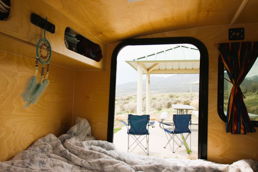Inside a cozy RV rental: soft bed, dream catcher, and stunning mountain campsite views.