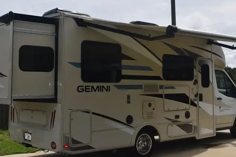 This light-colored Gemini RV rental, with its slide-out extended and awning deployed, is parked on a paved surface, ready for travel.