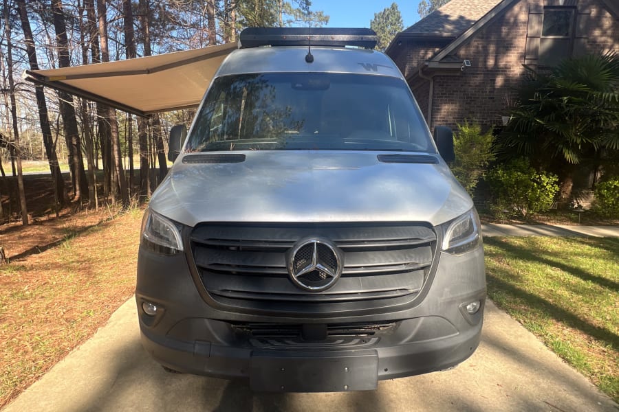 Front view of a silver Mercedes Sprinter RV rental parked on a sunny driveway, with an extended awning and trees nearby.