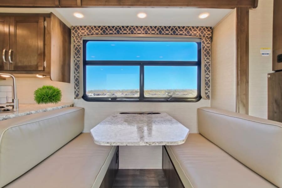 Bright RV rental interior featuring a spacious dinette with bench seating, table, and a large window. Kitchenette visible on left.