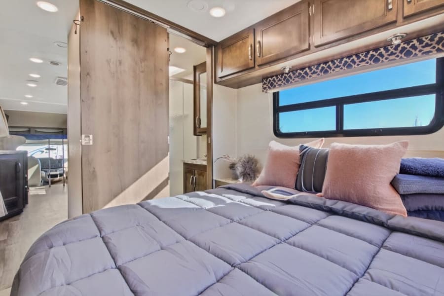 Modern RV rental bedroom: cozy bed, overhead cabinets, window with blue sky, and sliding door to private bath. Travel comfort.