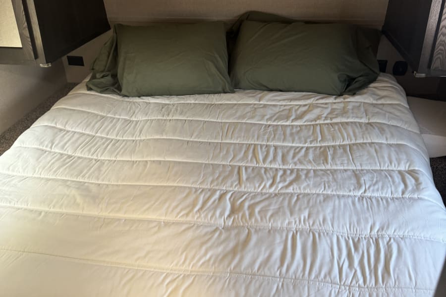 Inside an RV rental: A neat bed with a white quilted comforter and olive pillows, showcasing the cozy sleeping area.