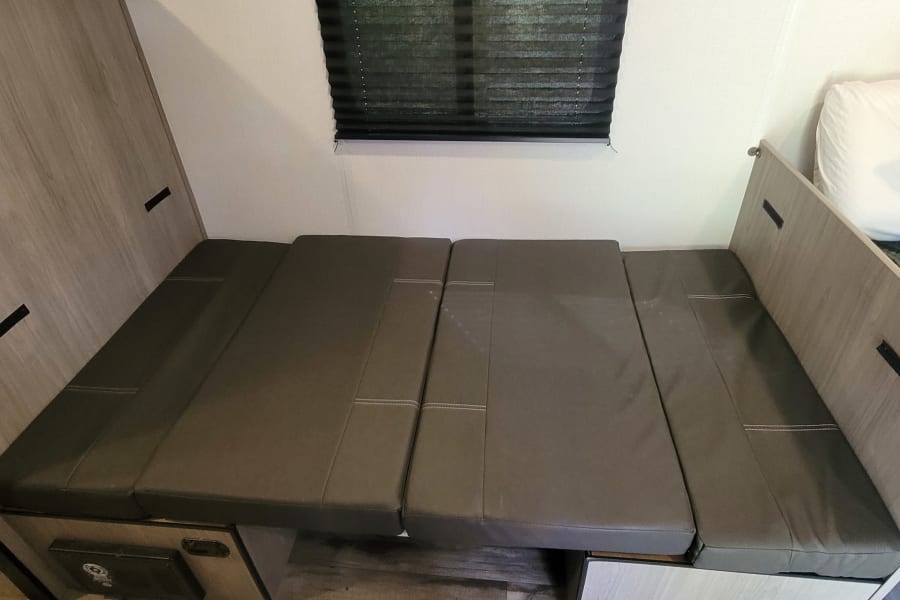 RV rental interior: dinette converted into a bed with gray cushions and a window blind.