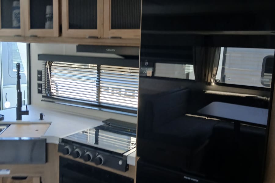 Modern RV rental kitchen with Furrion refrigerator, stove, microwave, and light wood cabinets. Perfect for travel.