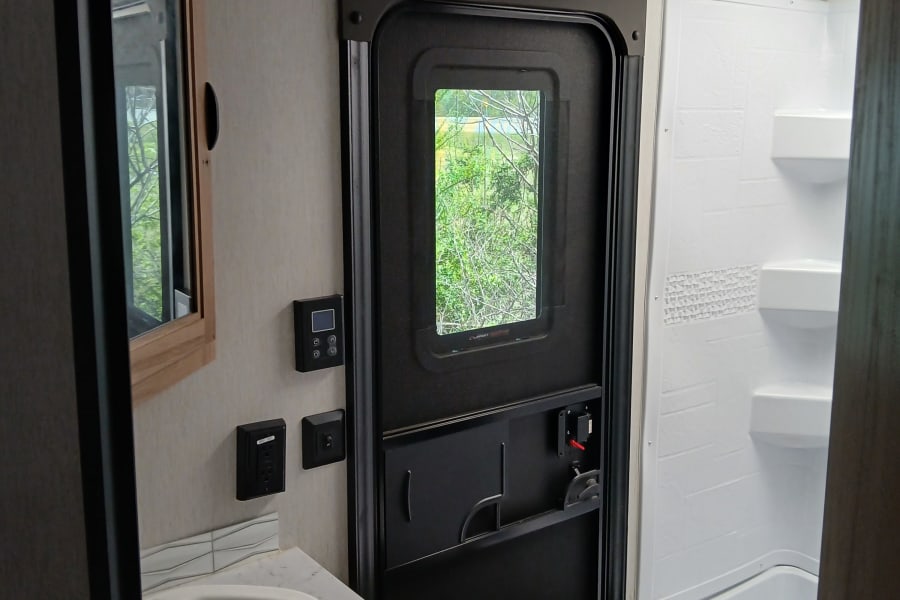 Inside an RV rental, showing a compact bathroom with a sink, shower, toilet, and main entry door with a window.
