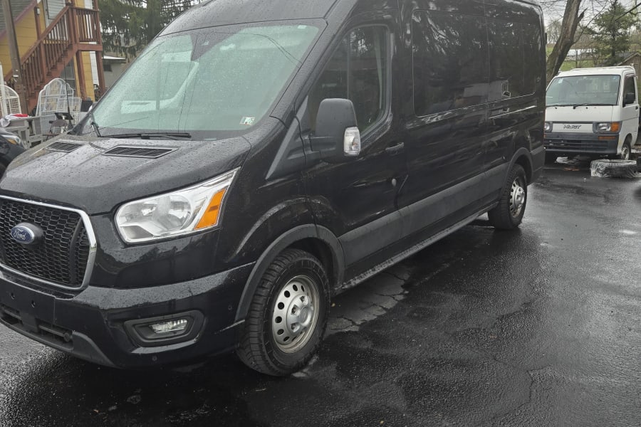 A black Ford Transit van, commonly used for RV rental conversions or commercial transport, sits on wet pavement. Another white van is behind.