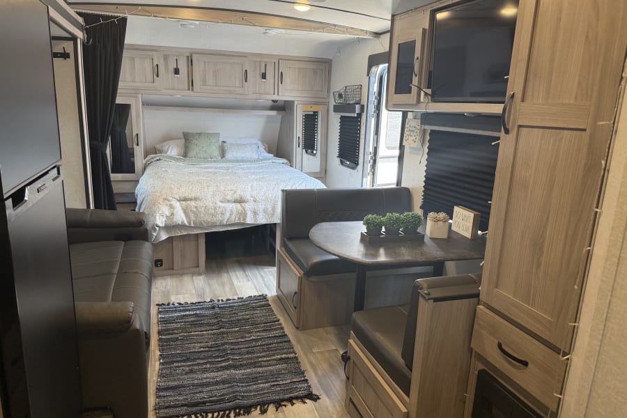 Bright rv rental interior showcases a cozy bed, comfortable dinette, sofa, and TV, with light wood finishes.