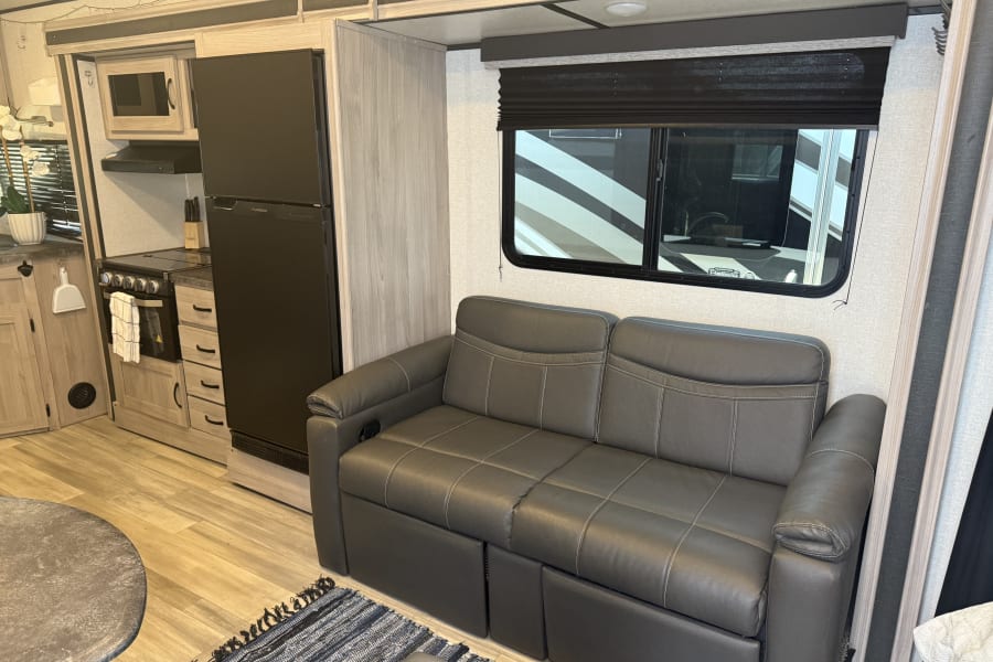 RV rental interior showcasing a kitchen with fridge & oven, and a dark grey reclining sofa.