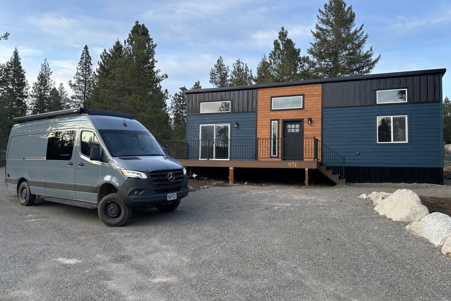 Gray Sprinter van (rv rental) next to a sleek blue & wood tiny house, amidst tall pines.