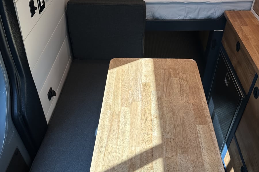 Bright RV rental interior showing a versatile dinette table, bench seat, and elevated bed area.