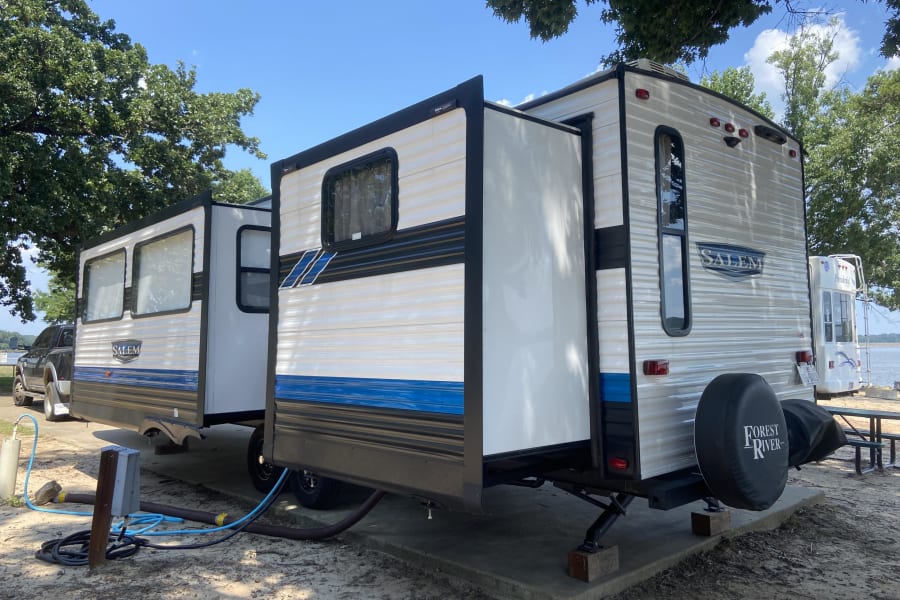 Experience RV rental life! A spacious travel trailer with extended slide-outs set up at a lakeside campsite with full hookups.