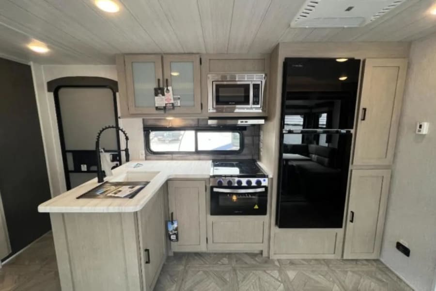 RV rental interior: functional kitchen with modern black fridge, microwave, stove, sink, and light wood cabinetry.