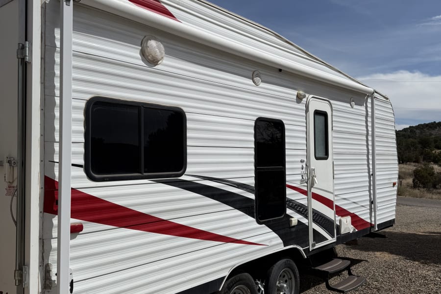 An RV rental with steps and a blue/white rug, parked on gravel under a blue sky. Features red & black stripes.