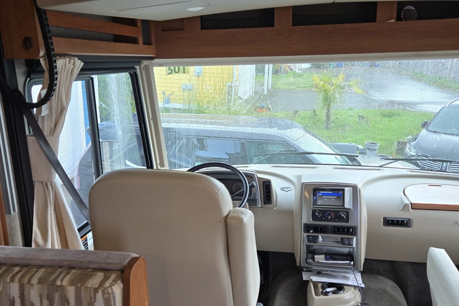 Inside this RV rental: See the dashboard, driver's area, and overhead bunk. Outside, a rainy day unfolds through the windshield.