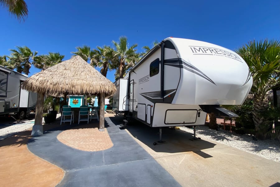 Tropical RV rental at a sunny park, featuring a large fifth-wheel RV and a charming tiki hut with seating. Perfect vacation spot.
