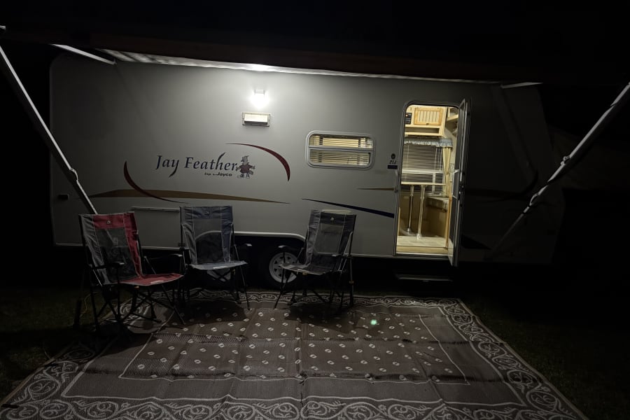 Nighttime RV rental setup: Illuminated Jay Feather travel trailer with open door, extended awning, camping chairs, and outdoor mat.