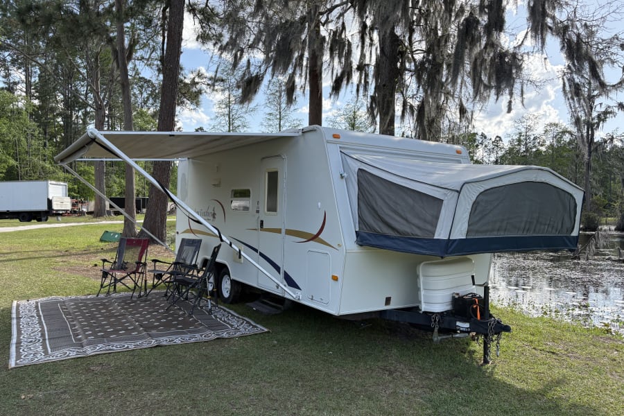 RV rental at a scenic campsite. Features a white travel trailer with awning, chairs, and a lake view, perfect for outdoor adventures.