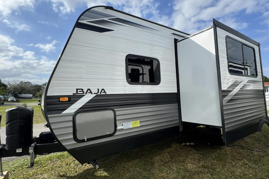 Modern RV rental travel trailer, a Baja model with extended slide-out, parked on grass.