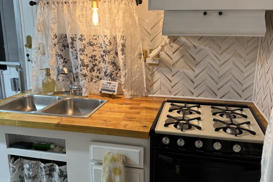 Renovated RV rental kitchen with double sink, wood counter, stove, microwave, white cabinets, and modern backsplash.