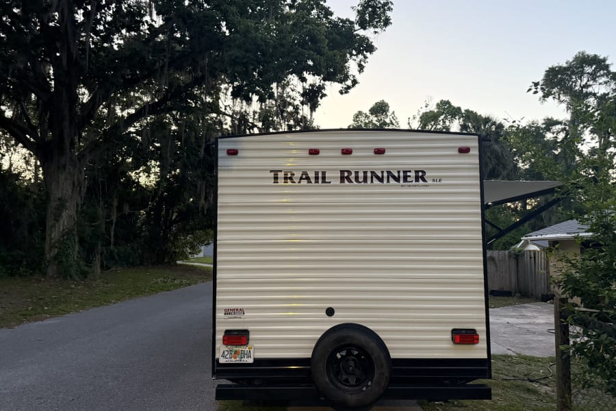 Rear view of a Trail Runner RV rental, a travel trailer by Heartland, parked on a paved drive near trees.
