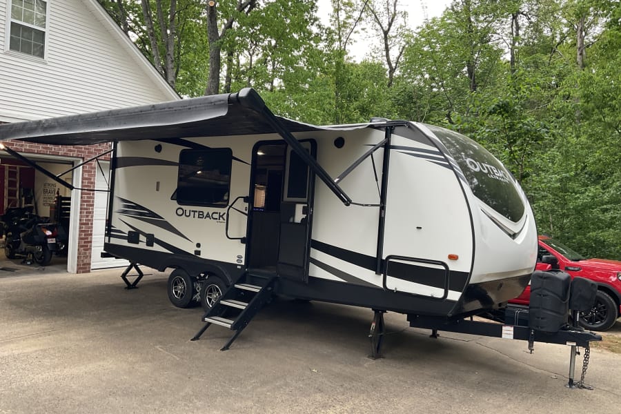 Outback travel trailer, white with black accents, ready for an RV rental. Awning extended, steps down, parked on a driveway.