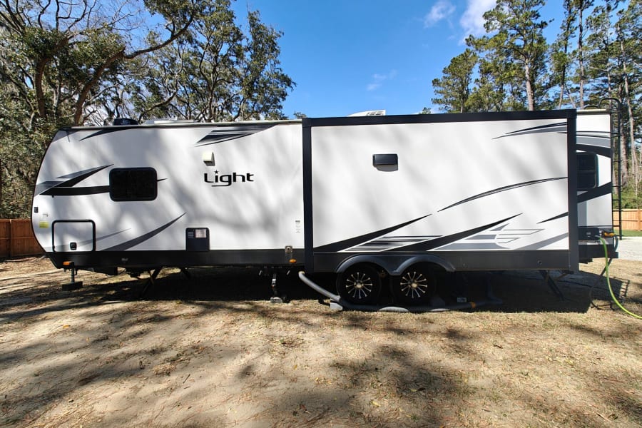 White "Light" travel trailer RV for rv rental, parked and hooked up at a sunny treed campsite.