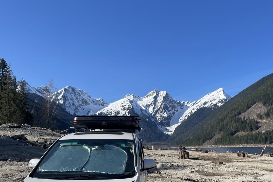 White RV rental van facing snow-capped mountains, a lake, and a dry bed. Scenic BC outdoor adventure awaits!