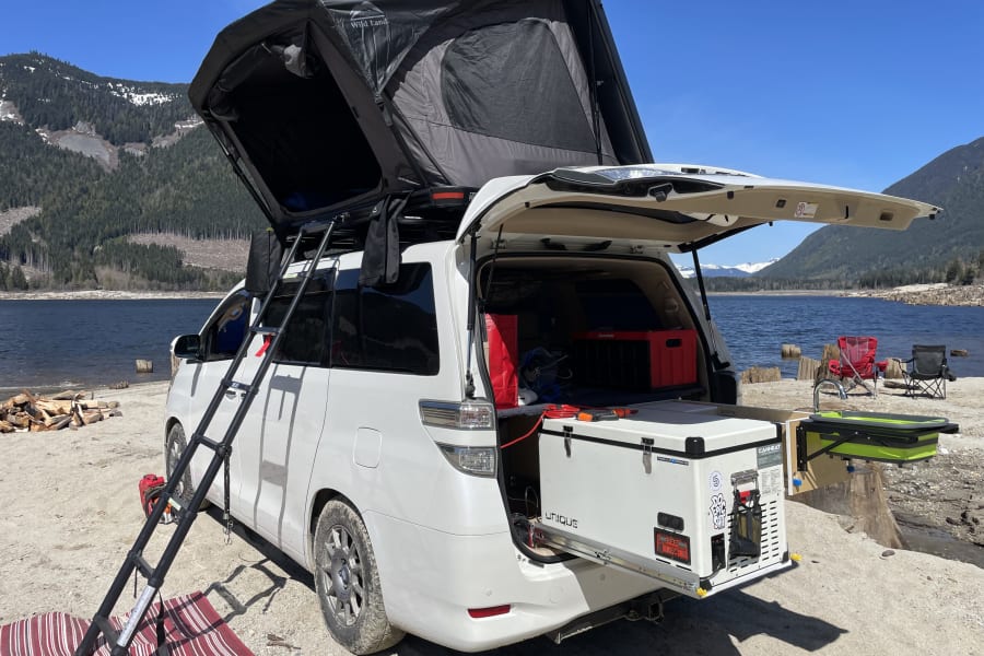 Ultimate RV rental setup: white camper van with deployed rooftop tent, ready for lakeside mountain camping.