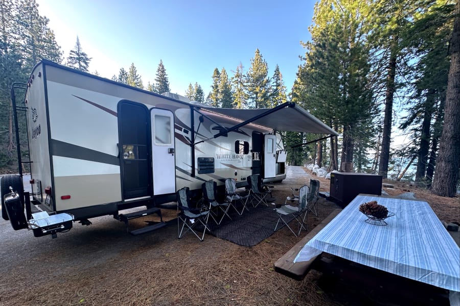 An RV rental travel trailer at a serene campsite with an open awning, outdoor seating, and a picnic table among tall pines.