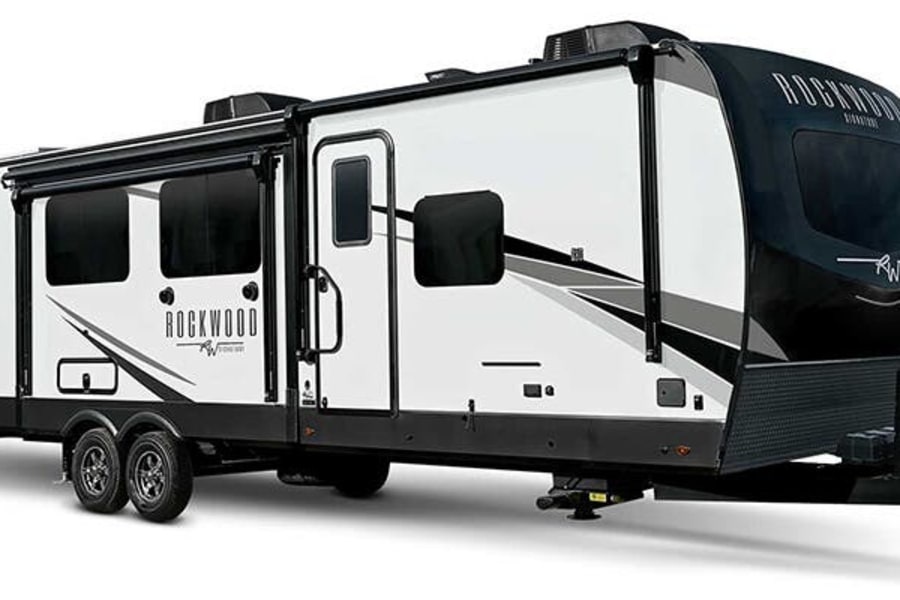 White and black Rockwood travel trailer, an ideal RV rental. This modern recreational vehicle features windows, a door, and a front hitch.