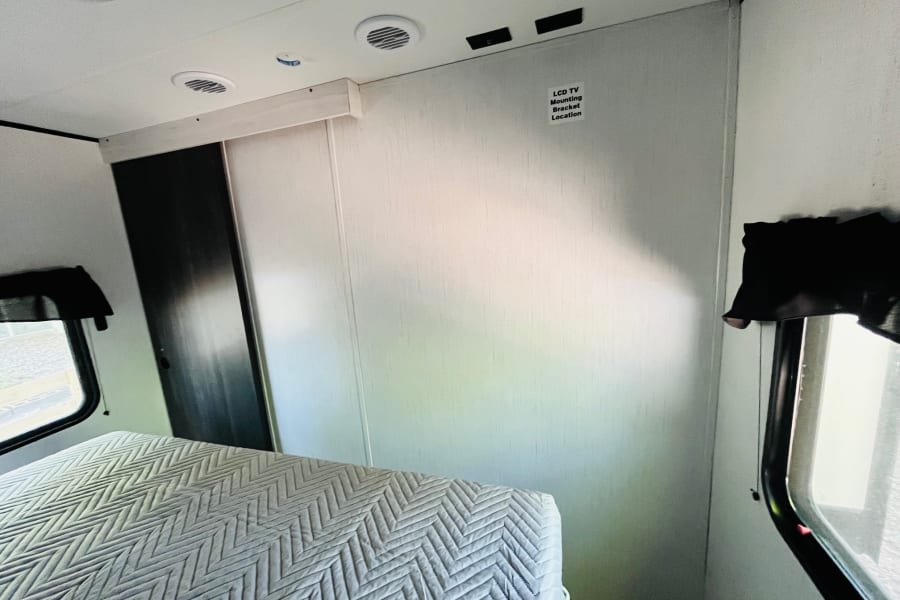 Inside an RV rental: a bright bedroom with a quilted bed, windows, and a dedicated wall for a TV mount.