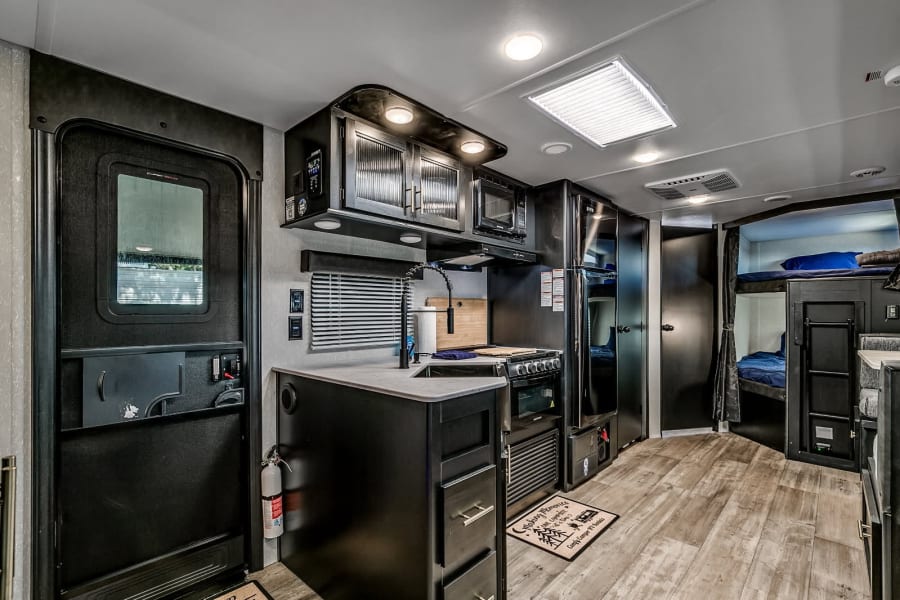 This RV rental interior features a dark modern kitchen, complete with a sink, microwave, and stove, plus bunk beds.