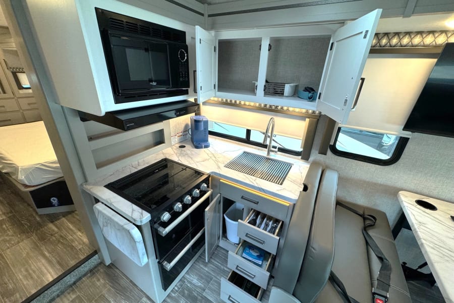 Inside a modern RV rental: a compact kitchen with a microwave, stove, sink, pull-out drawers for utensils, and a comfortable dinette.
