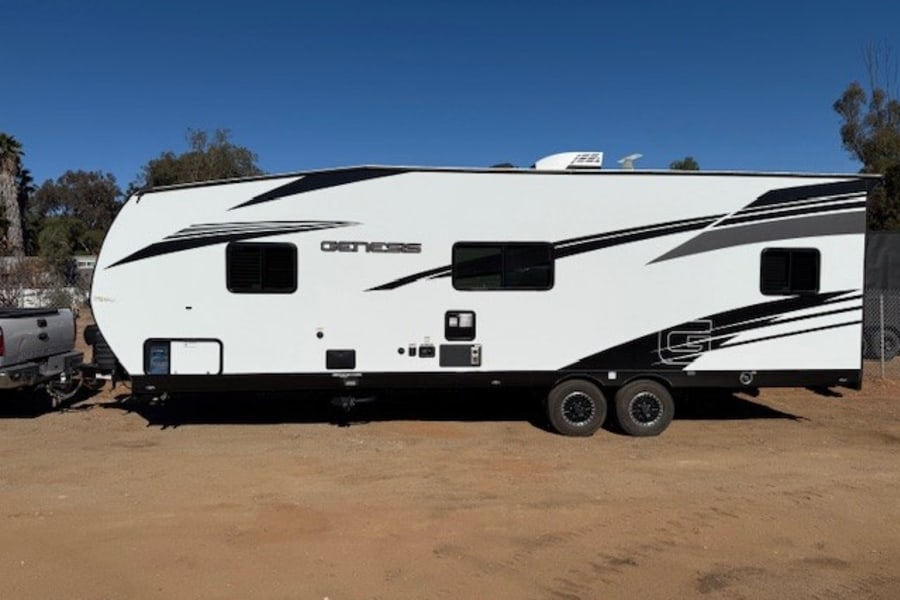 White Genesis travel trailer RV rental parked on dirt. This sleek unit features black & grey graphics & multiple windows.