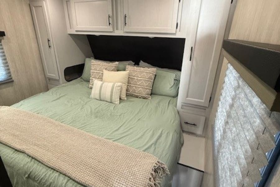 Cozy RV rental bedroom: light green bed, ample overhead/side storage, and a bright window with blinds. Perfect for travels.