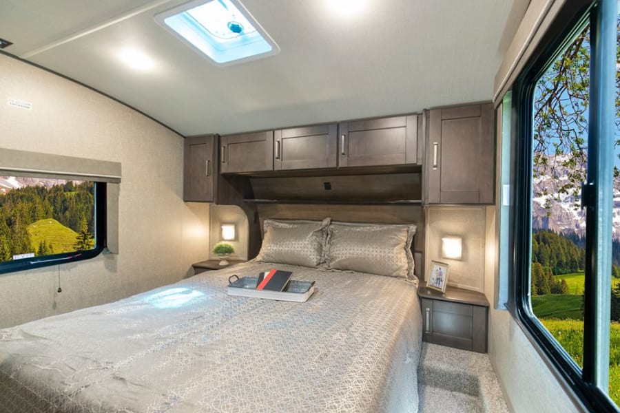 Spacious RV rental bedroom featuring a large bed, overhead cabinets, and dual windows showcasing stunning natural views.
