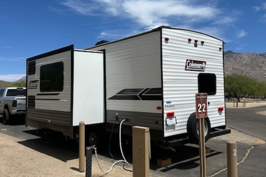 White & grey travel trailer RV rental parked at a sunny campsite with hookups. Site 22, ready for your next adventure.