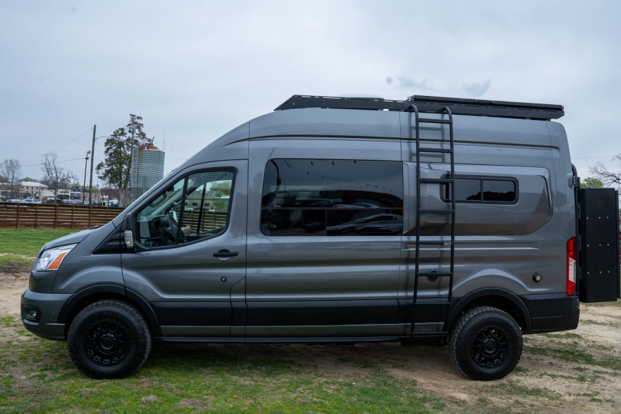 A custom gray Ford Transit camper van, perfect for RV rental adventures, with a roof rack, side ladder, and all-terrain tires.