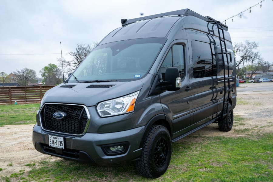 A dark gray Ford Transit van converted into a camper, equipped with a roof rack and ladder. Ideal for RV rental.