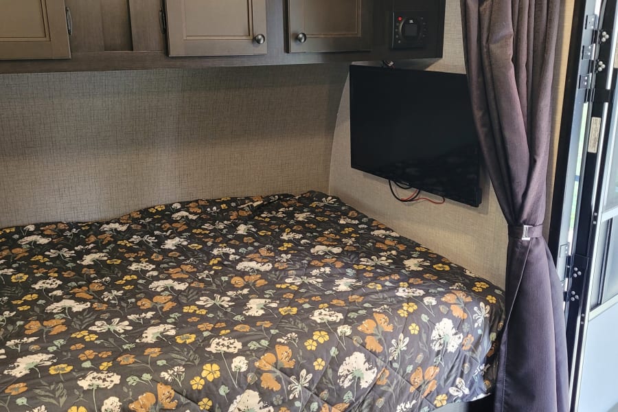 RV rental interior: cozy bedroom with floral bed, overhead cabinets, mounted TV, and under-bed storage. Compact comfort.