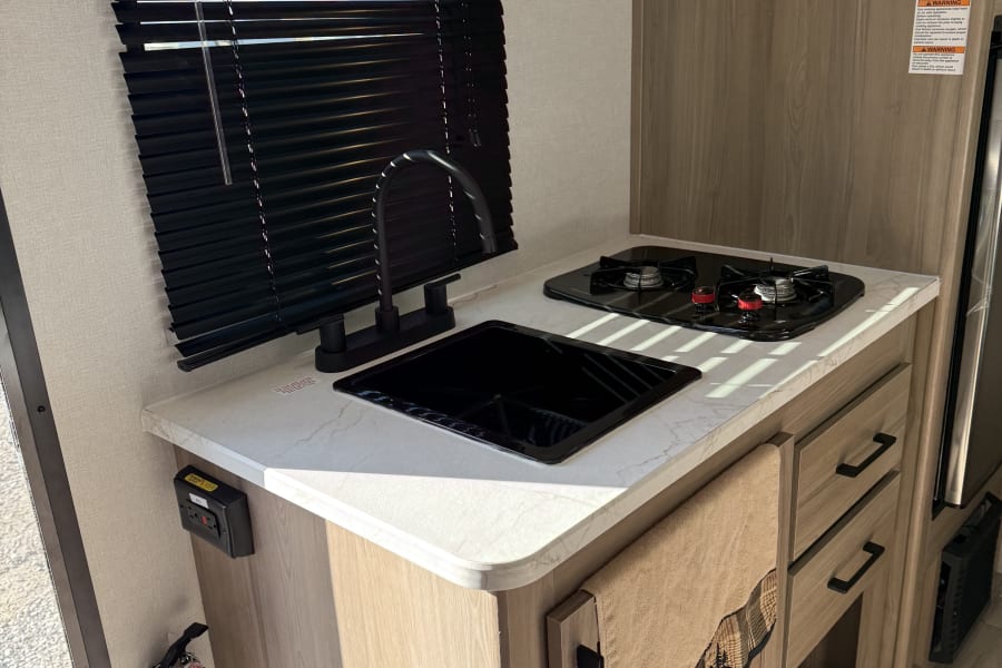 RV rental kitchen interior: functional galley featuring a modern sink, stove, light wood cabinets, and window blinds.